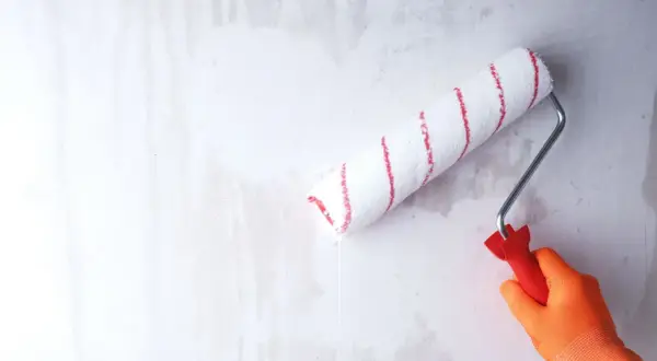 Ręka w pomarańczowej rękawicy wykonuje gruntowanie przed malowaniem, używając wałka z czerwonymi paskami.