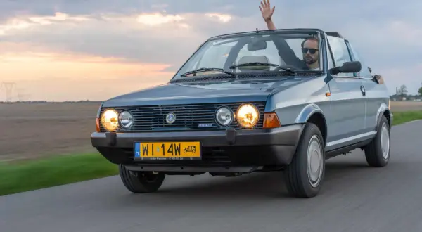 Mężczyzna w okularach przeciwsłonecznych macha ręką z kabrioletu Fiat Ritmo. Jazda po drodze o zachodzie słońca.