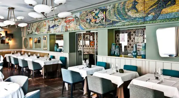 Interni eleganti di un ristorante, con tavoli apparecchiati e un grande mosaico marino sul muro. Un luogo ideale per scoprire i migliori ristoranti di pesce a Milano.