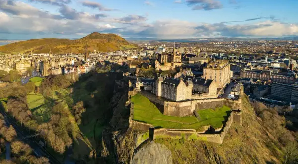 Edinburgh Castle tronar över staden, Skottlands huvudstad. En historisk vy med gröna kullar och moln på himlen.