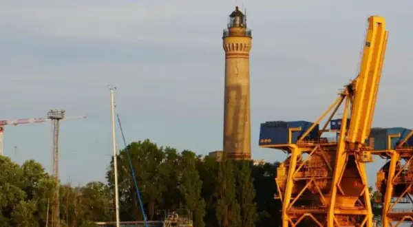 Wysoka, żółta latarnia morska w Gąskach góruje nad portem, obok potężnych, żółtych dźwigów portowych i zielonych drzew.
