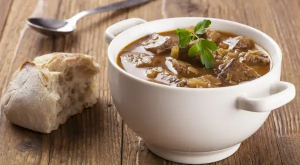 Aromaticzny, tradycyjny gulasz wołowy w białej miseczce, udekorowany natką pietruszki, obok kromka chleba i łyżka.