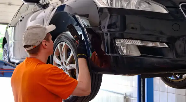 Mechanik w czapce i pomarańczowej koszulce ogląda podniesiony czarny samochód.