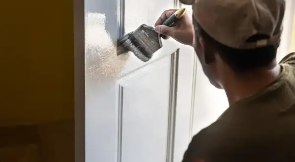 Mężczyzna w czapce z daszkiem maluje białe drzwi pędzlem. Odświeża wygląd wnętrza.