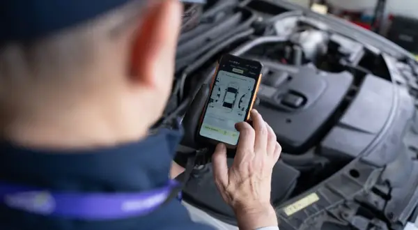 Mechanik w rękawiczkach trzyma smartfon z aplikacją do oceny stanu samochodu, na tle otwartej maski silnika.