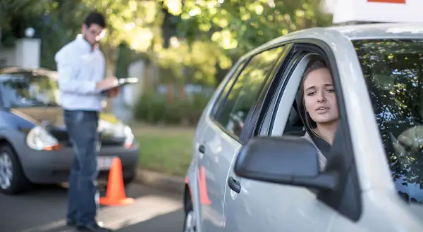 Joven nerviosa al volante, con un instructor anotando. ¿Se puede sacar el carnet de conducir sin autoescuela?