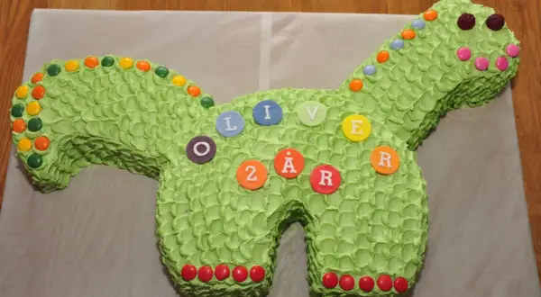 Enkel dinosaurietårta i ljusgrönt med godisdekorationer. Tårtan är formad som en dinosaurie och har bokstäver och siffror som bildar "OLIVER 2 ÅR".