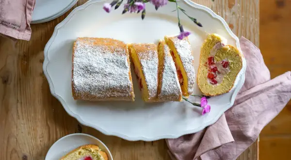Eine köstliche Erdbeer Biskuitrolle, mit Puderzucker bestäubt und mit frischen Erdbeeren gefüllt, liegt auf einer weißen Platte.