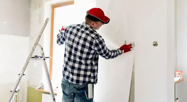 Mann mit roter Kappe und Handschuhen kleistert Vliestapete ein, um eine Wand zu tapezieren.
