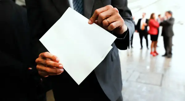 Mężczyzna w garniturze trzyma białą kopertę, jakby miał dokonać przekierowania. W tle ludzie w eleganckich strojach.