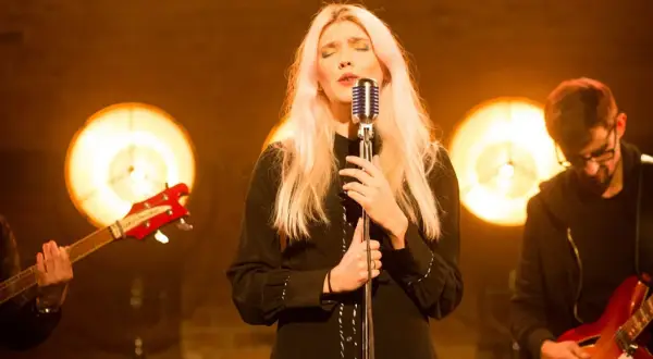 Wokalistka z blond włosami śpiewa przy mikrofonie, w tle zespół z gitarami i światła.