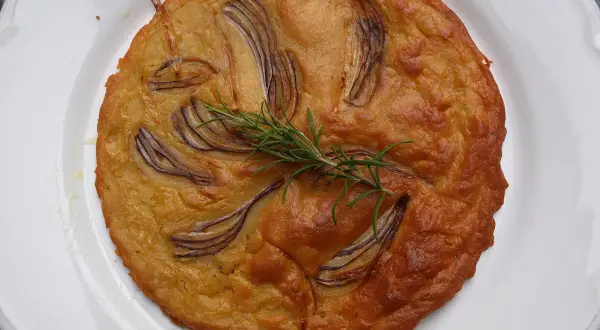 Herzhafte Kichererbsenmehl Rezepte: Goldbrauner Kuchen mit roten Zwiebeln und Rosmarin auf weißem Teller.