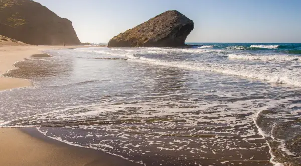 La duna de Monsul se extiende hacia el mar, con olas rompiendo suavemente en la arena dorada. Una figura solitaria camina por la playa.
