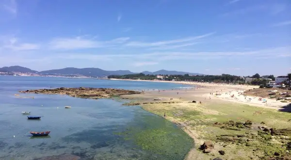 Vista de la playa de A Calzoa en Vigo, con gente disfrutando del sol y el mar. Barcas pequeñas flotan en aguas cristalinas.