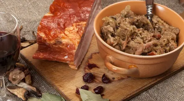 Ania gotuje bigos. Na desce kawałek wędzonego boczku, miseczka z bigosem, kieliszek czerwonego wina, suszone grzyby i liście laurowe.