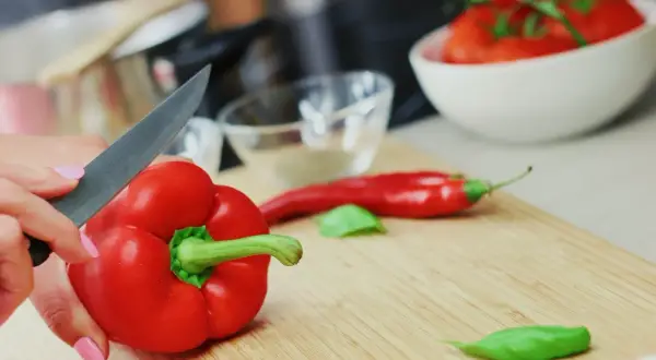 Ręka kroi paprykę czerwoną, bogatą w witaminy. Obok leży chili i pomidory, składniki pełne zdrowotnych właściwości.