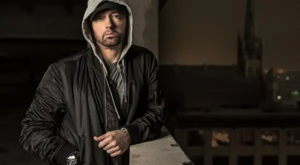 Eminem w czarnej kurtce bomberce i szarej bluzie z kapturem, oparty o betonową barierkę.