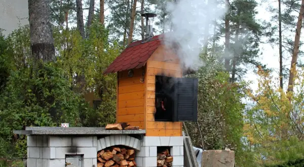 Gotowa budowa wędzarni ogrodowej. Dym unosi się z komina, a w środku wiszą kiełbaski.
