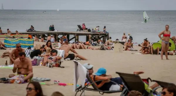 Tłum ludzi na plaży, opalanie się i relaks. Idealna pogoda na lipiec nad morzem, widać żaglówkę i wiatrak.