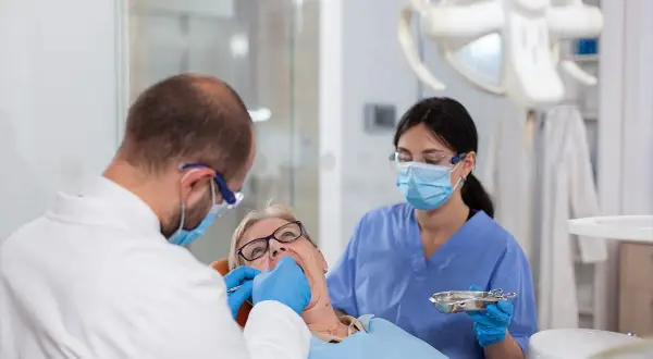 Dentysta zakłada białe plomby na NFZ starszej pacjentce. Asystentka stomatologiczna pomaga.