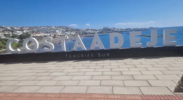 Napis "COSTA ADEJE TENERIFE SUR" na tle oceanu i miasta. Uważaj na słońce i tłumy, by w pełni cieszyć się wakacjami na Teneryfie.
