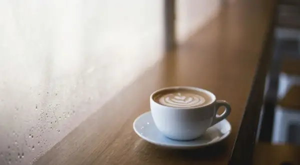Tazza di cappuccino con latte art su un tavolino di legno, vicino a una finestra appannata. Immagina come mettere le cialde nel portafiltro per un caffè perfetto.