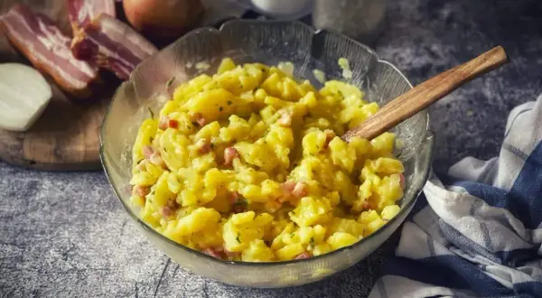 Ein dampfender, warmer Kartoffelsalat mit Speck und Zwiebeln, serviert in einer Glasschüssel mit Holzlöffel.