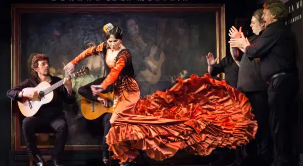 Una bailaora de flamenco, con un vestido naranja voluminoso, baila al son de guitarras. Es un espectáculo de flamenco vibrante.