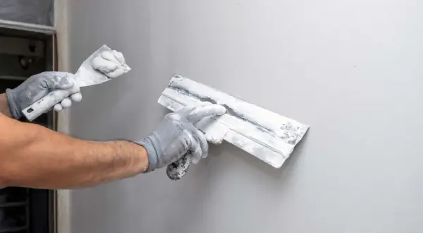 Mani guantate eseguono la rasatura di un muro con spatole cariche di stucco, preparando la superficie per la finitura.