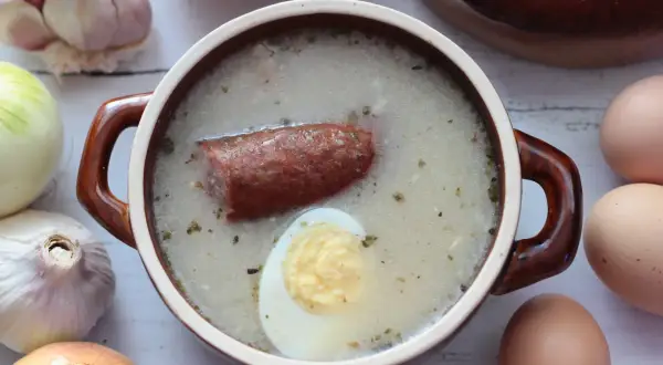Gotowy zakwas na żurek śląski rączka gotuje. W garnuszku pływa kiełbasa i jajko. Obok cebula, czosnek i jajka.