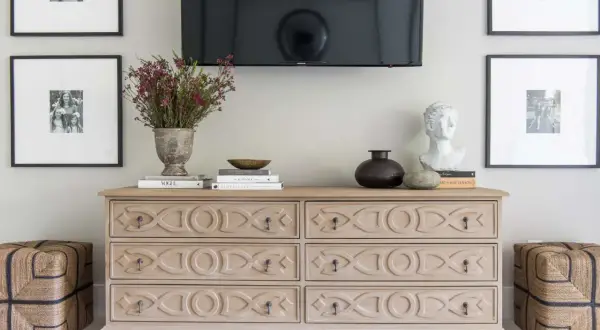Ideas on how to decorate a dresser: a light wood dresser with decorative carvings, topped with books, a vase of flowers, a bust, and a dark vase.