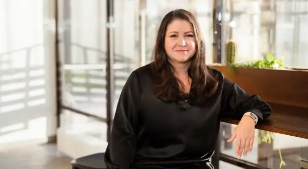 Aida Alvinius, en kvinna med långt brunt hår, ler mot kameran. Hon bär en svart klänning och sitter vid ett träbord med växter i bakgrunden.