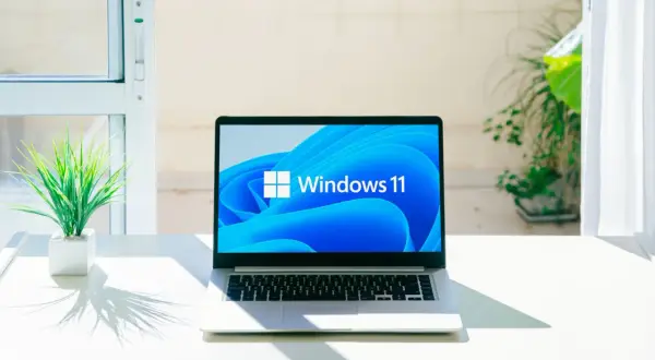 Laptop z logo Windows 11, gotowy na aktualizację 25H2. Na biurku obok rośliny, w jasnym pomieszczeniu.