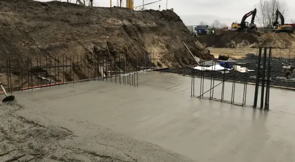 Budowa w toku: świeża wylewka betonowa na zewnątrz, zbrojenie i koparki w tle.