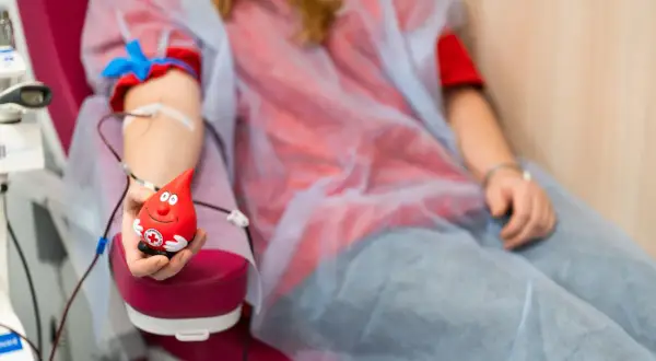 Uśmiechnięta kropla krwi trzymająca zabawkę, symbol tego, czy oddawanie krwi jest zdrowe. Osoba w czerwonej koszulce i niebieskim fartuchu oddaje krew.