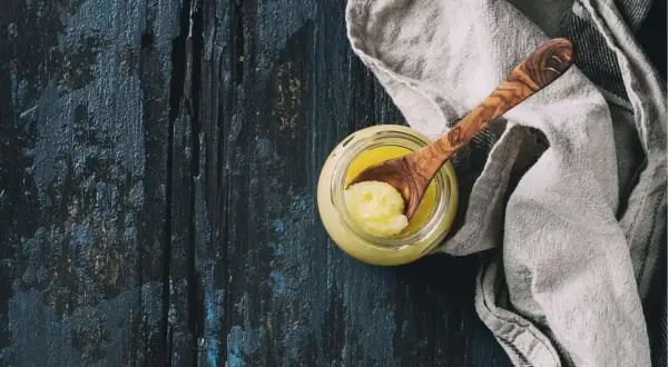 Ein Glas mit goldgelbem Ghee, daneben ein Holzlöffel mit einer Portion. Ghee ist geklärte Butter, ein vielseitiges Kochfett.