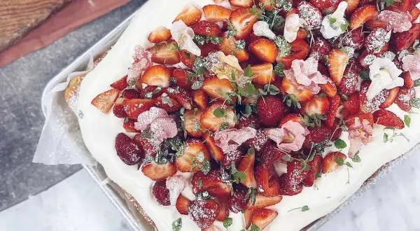 En midsommartårta i långpanna, täckt med fluffig grädde och generöst toppad med färska jordgubbar och små rosa blommor. Perfekt för sommarens firanden!