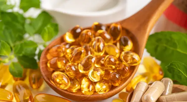 Łyżka pełna kapsułek z płynnym złotem – to suplementacja omega 3. Obok zielone liście i inne suplementy.