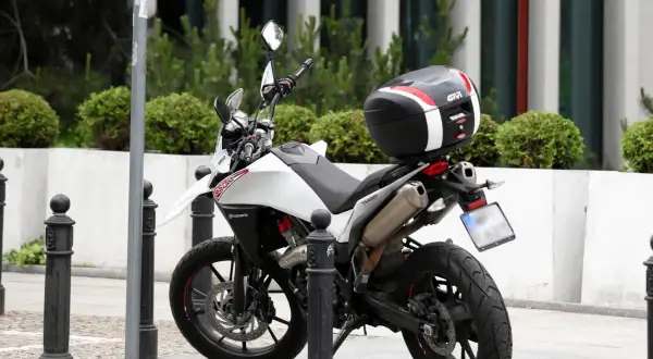 Biały motocykl z czarnym kufrem Givi zaparkowany na chodniku, obok słupków.