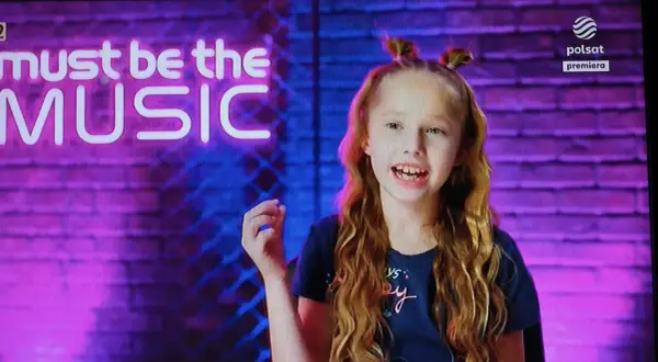 Lili chce śpiewać autor. Dziewczynka z burzą rudych loków i dwoma kucykami na głowie, na tle neonowego napisu "must be the MUSIC".
