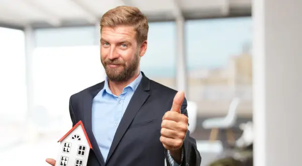 Ein Mann im Anzug hält ein Hausmodell und zeigt Daumen hoch. Er ist ein Investor, der in Immobilien investiert.