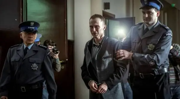 Scena z filmu o Tomaszu Komendzie: policjanci eskortują go w kajdankach, a za nimi stoi fotograf z aparatem.