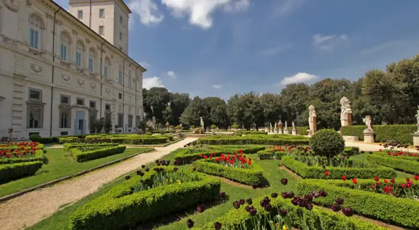 Giardini all'italiana di Villa Borghese con tulipani rossi e viola, statue e un palazzo sullo sfondo.