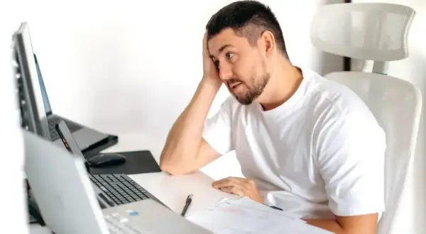 Zamyślony mężczyzna przy biurku z laptopami i dokumentami, zastanawia się, czy umowa zlecenie wlicza się do stażu pracy.