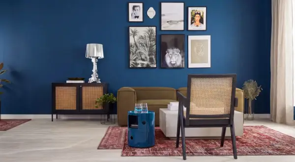 Ein stilvolles Wohnzimmer mit einer tiefblauen Wand, die mit Kunstwerken geschmückt ist. Ein Rattan-Stuhl steht vor einem Sofa, daneben ein runder, blauer Beistelltisch.