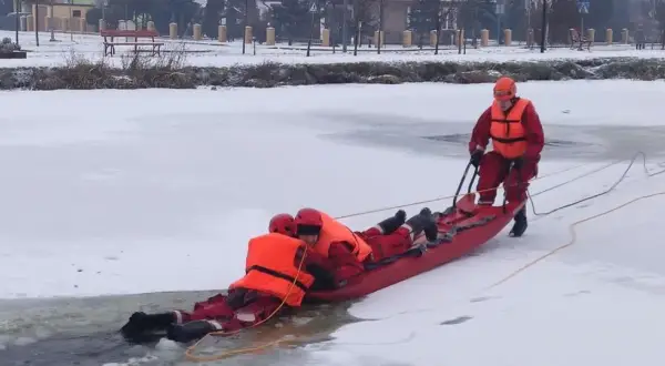 Ratownicy w akcji, wyciągają osobę z lodowatej wody za pomocą **sanki ratownicze**. Zimowa sceneria i profesjonalny sprzęt.