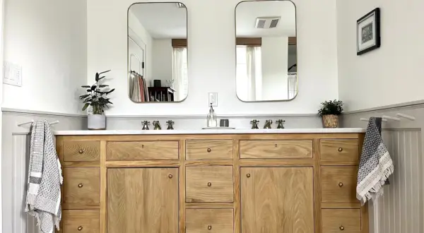 A beautiful DIY bathroom vanity with a natural wood finish, marble countertop, and brass fixtures. Two mirrors hang above, reflecting the bright, airy space.