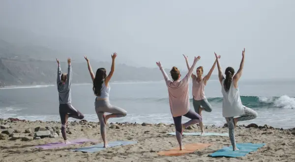 Cinco mujeres practican poses de yoga para 4 en la playa, con los brazos levantados hacia el cielo.
