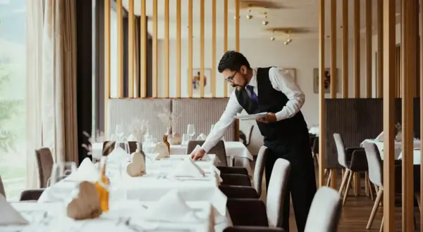 Cameriere in elegante divisa prepara la sala per la cena, un esempio di vitto e alloggio curato nei dettagli.