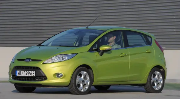 Zielony Ford Fiesta Mk7 z widocznymi żarówkami z przodu, zaparkowany przed budynkiem.